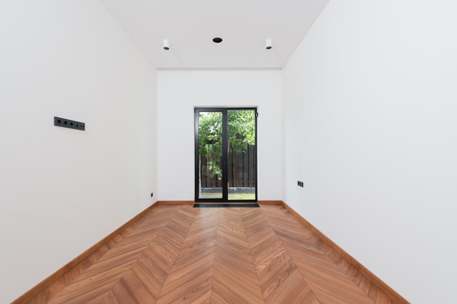 A clean, minimalist interior room with white walls, wooden parquet flooring, and a glass door.